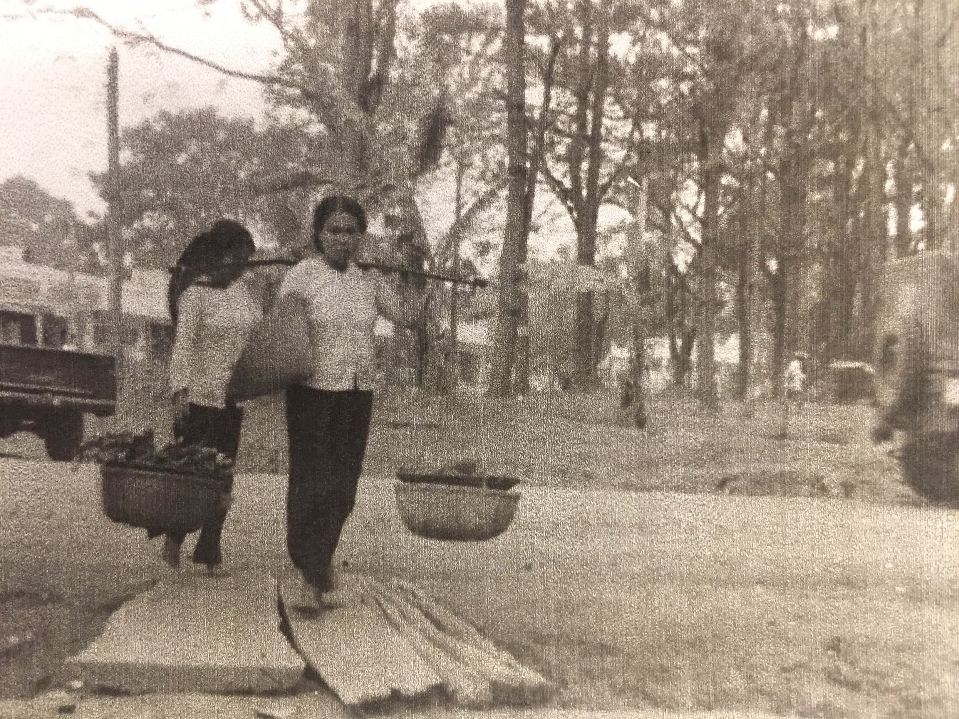 Two older Asian women on the street, one carrying a yoked basket full of food.