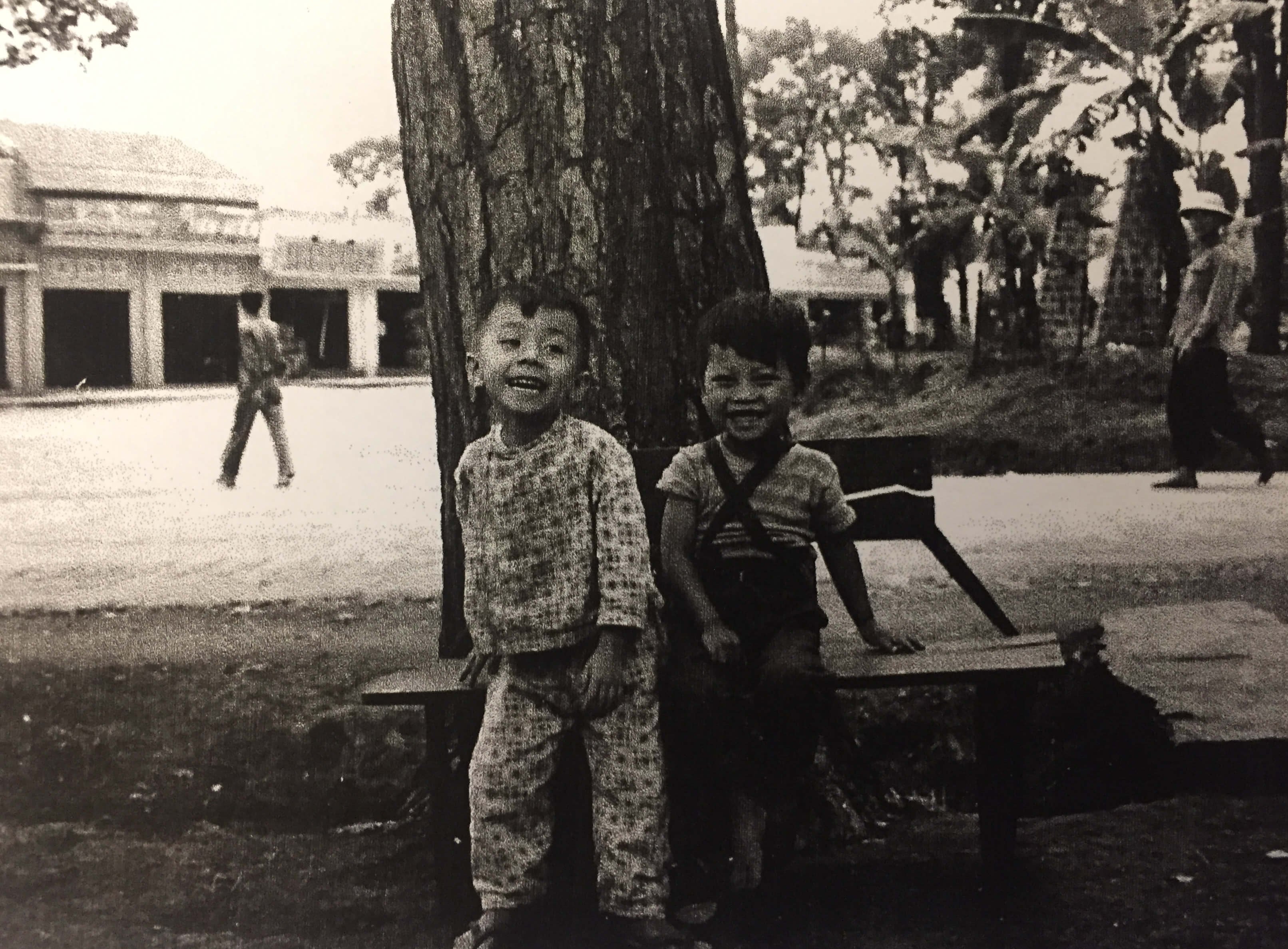 Two young Asian boys laughing on a bench under the shade of a tree.