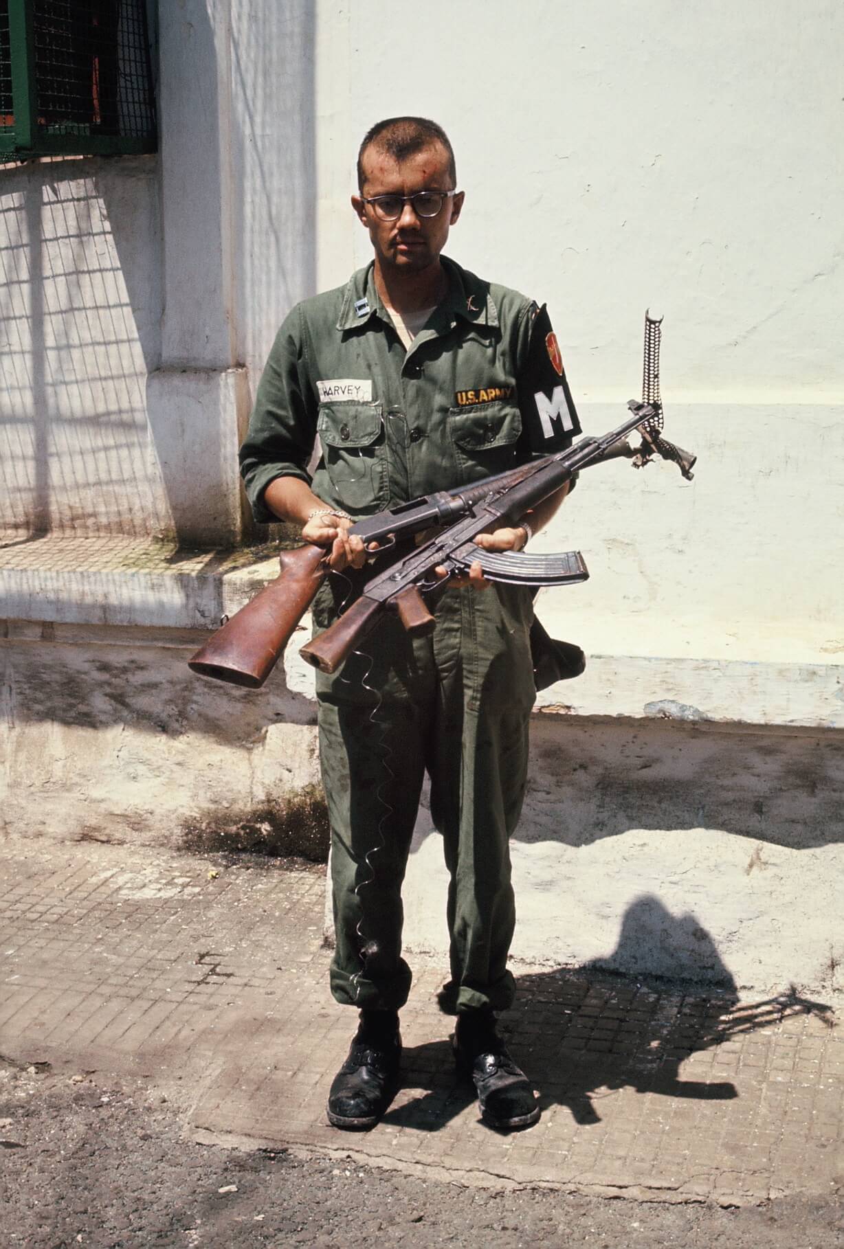 Military police officer holding two guns, standing out on the street.