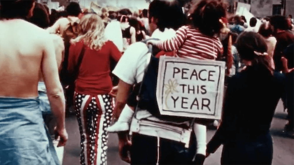 Protesters in the street. A child is strapped to her fathers back with a sign that says "Peace this Year".