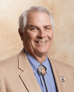 Contemporary portrait of an older gentleman in a blue shirt, tan jacket, and bolo tie.