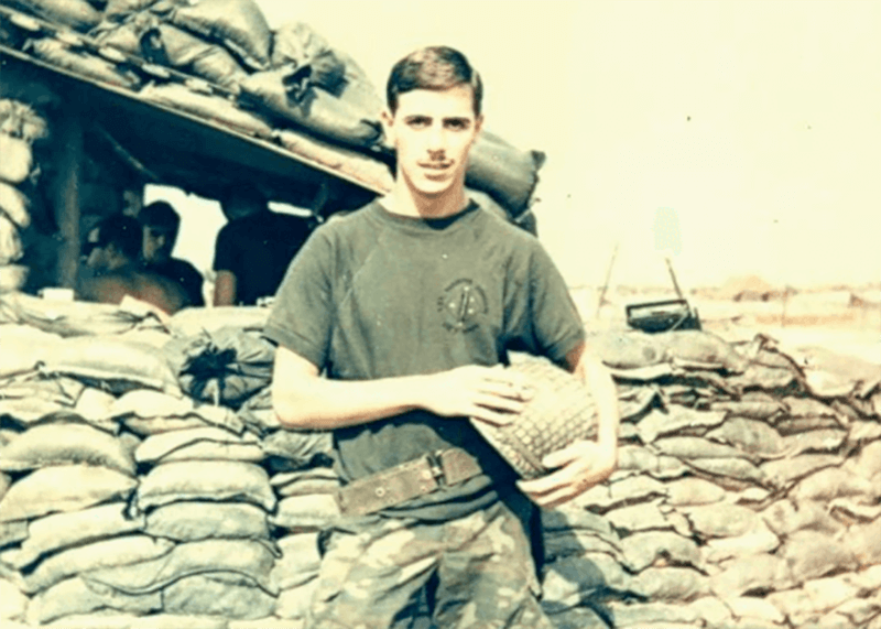 A young man with his helmet in his hands, standing in front of a sandbag bunker.