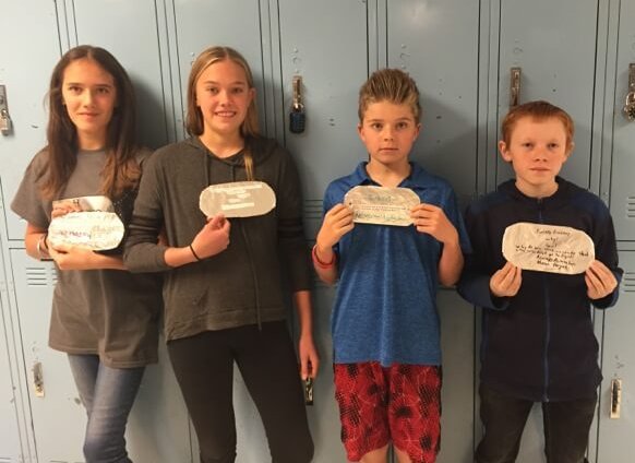 Contemporary photo of 2 middle school girls and 2 middle school boys posing in front of blue lockers and holding large "dog tag" school projects in front of them.