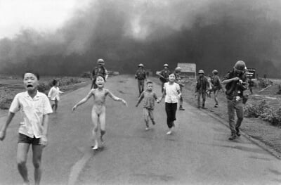 Group of soldiers and crying children, one naked, moving on foot away from a distant cloud of smoke rising from the ground.