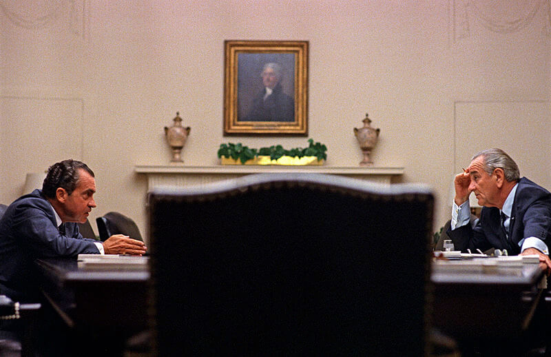 Nixon and Johnson looking intensely at one another across a long table. An empty office chair is in the foreground directly between them.