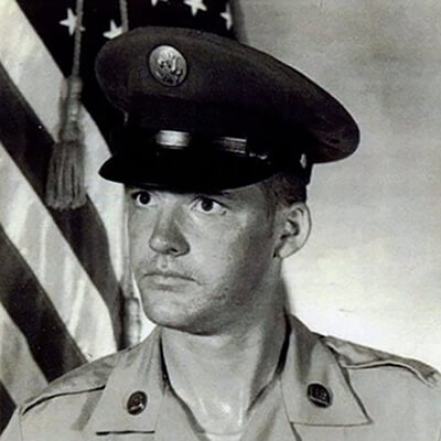 An official military portrait of a young man wearing a cap, looking stern, posed in front of an American flag.