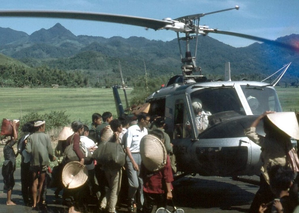 Asian civilians with bags and conical straw hats gathered around a helicopter with an American flag on it. Mountains in the background.