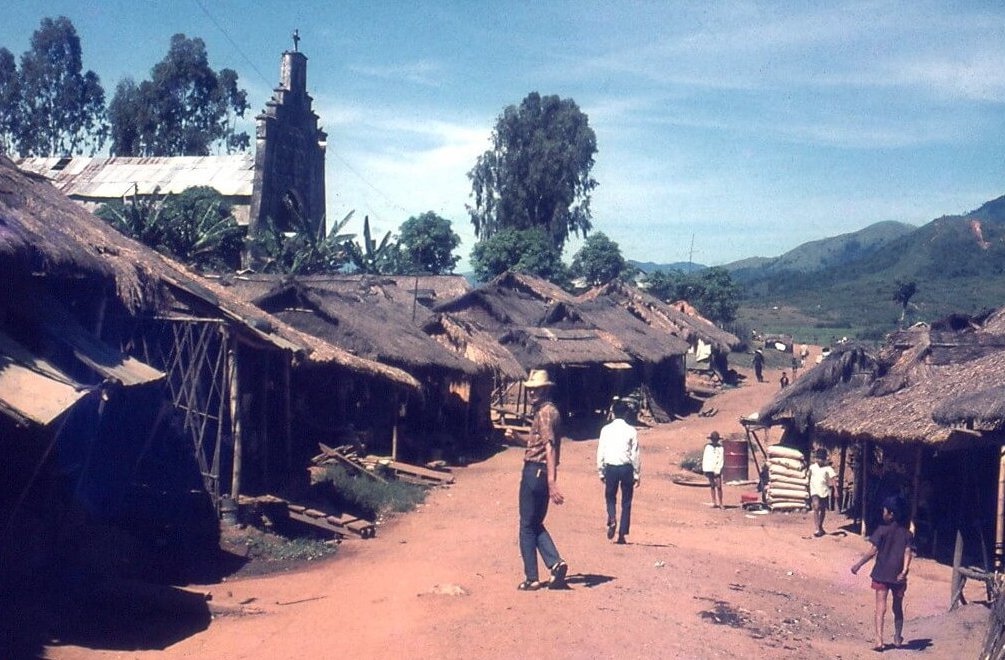A young American man in civilian clothes looking over his shoulder as he walks down a dirt path between thatched huts.