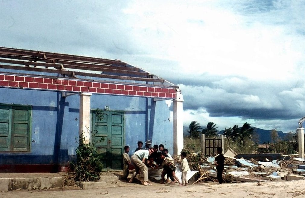 A young American man and several young Asian children crouching outside a blue, green, and red building.