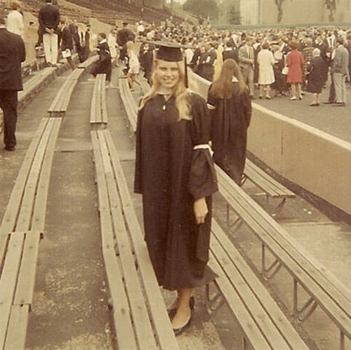 A young woman in graduation cap and gown, standing in emptied out bleachers.