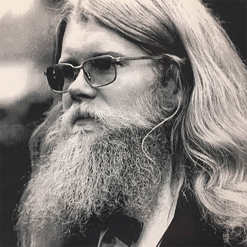 A man with long hair, a beard, and tinted glasses in a suit and bow tie with a carnation boutonniere.