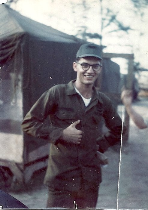 A young man in glasses and uniform, smiling and holding his right hand to his torso.