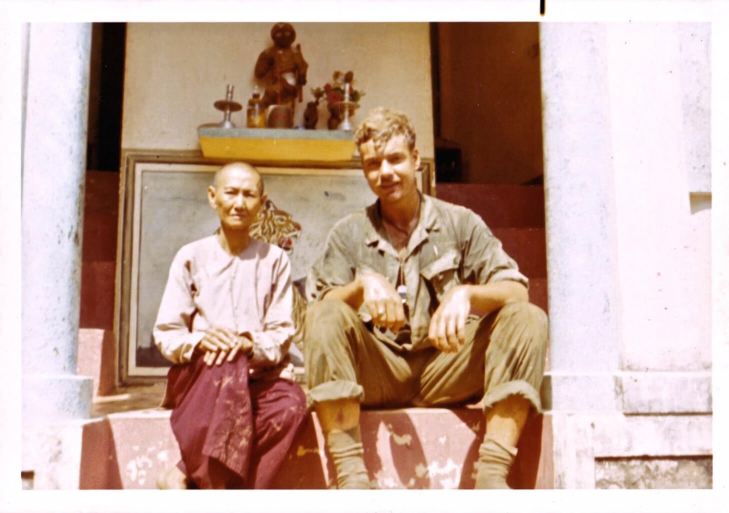 A young soldier sitting next to a smaller Asian man, on steps outside of a building.