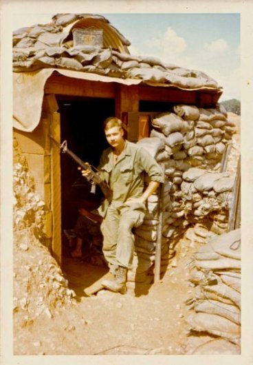 A young soldier with a gun propped on his leg, standing in the entrance of a bunker.