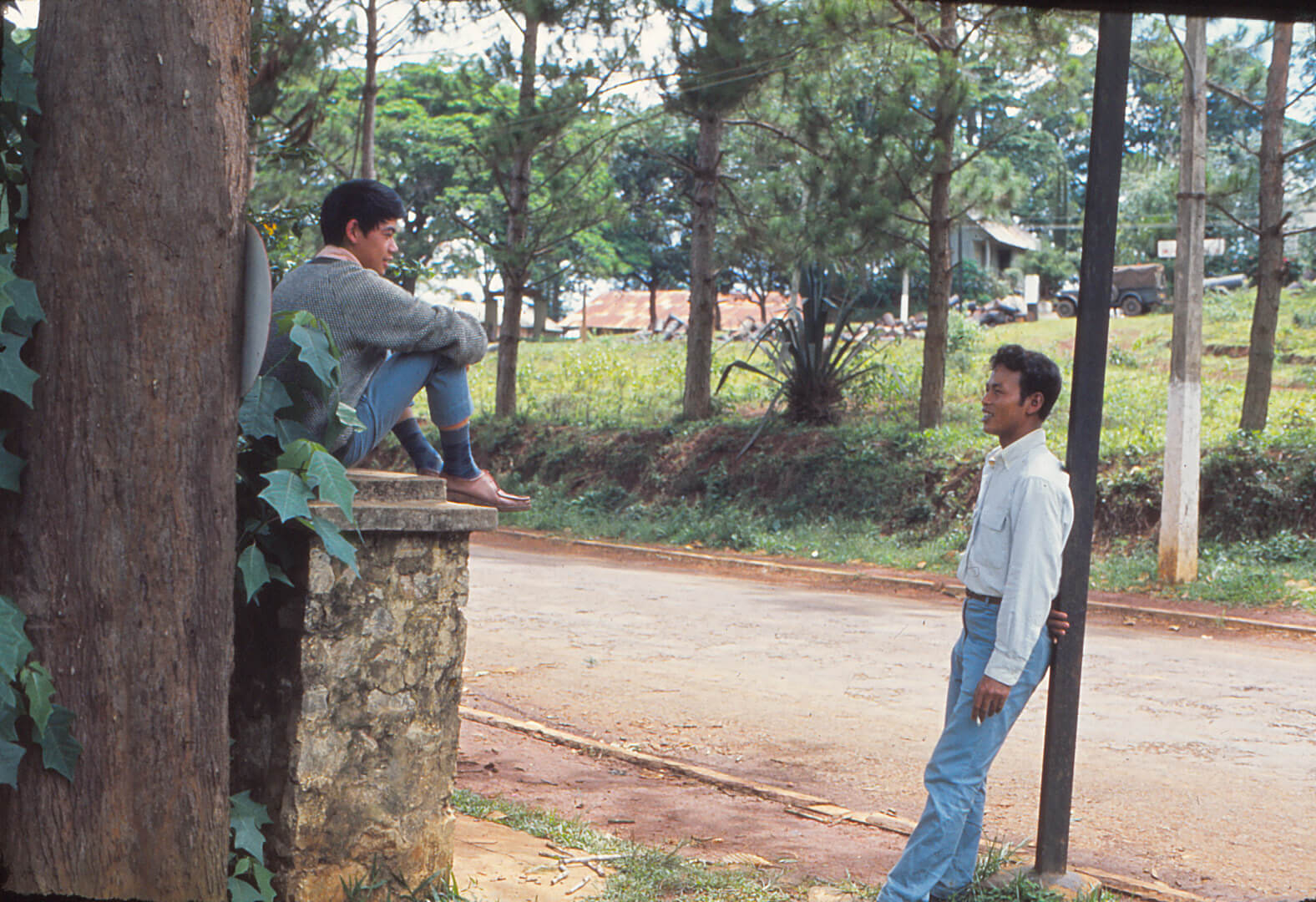 Two young Asian men, one leaning against a post with a cigarette, the other sitting casually atop a stone post.