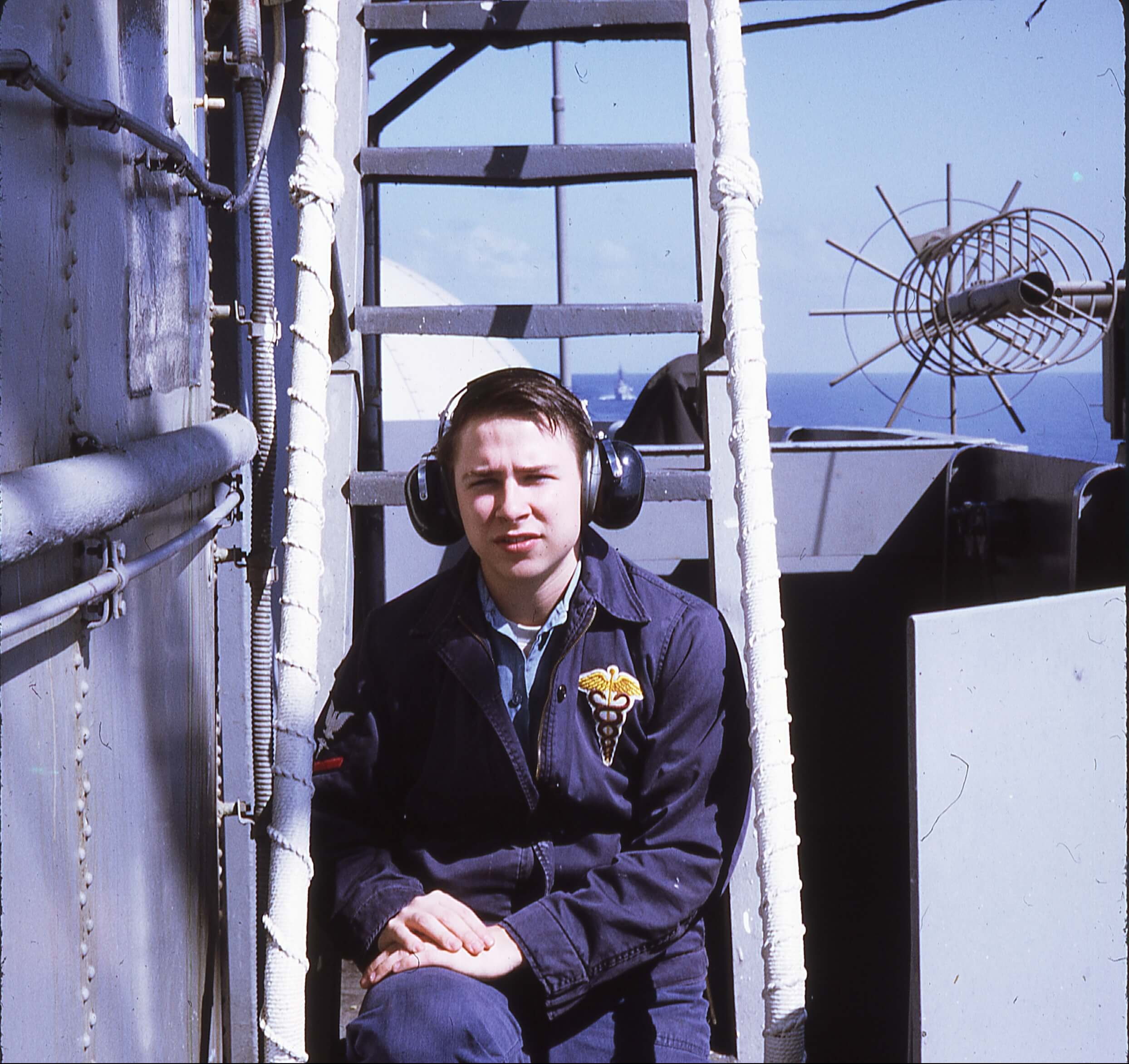 A young man in a navy blue jumpsuit wearing big headphones over his ears, sitting on a ladder on a boat.
