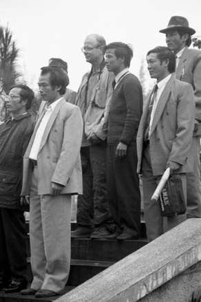 Black and white photo of Asian men in suits, and one white man in casual travel attire, standing on steps and looking off camera.