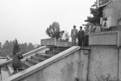 Black and white photo of a man taking the photo of several other men standing at the top of some steps, in front of a monument.