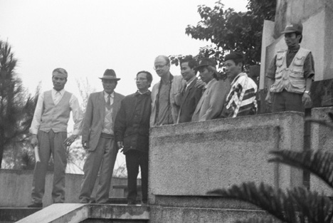 Black and white photo of Asian men in suits, and one white man in casual travel attire, standing on steps and posing for a photograph.