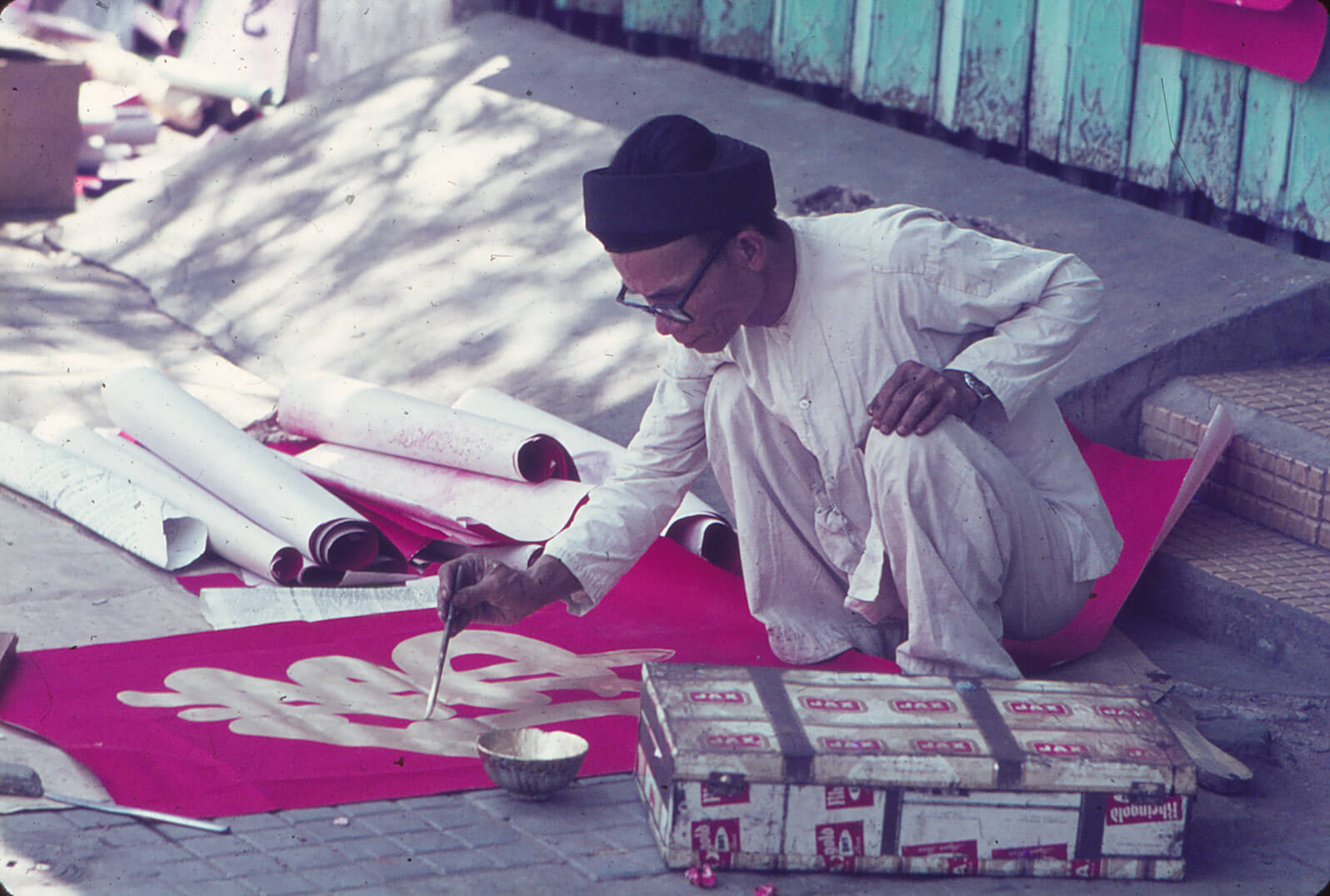 An older Vietnamese man in white clothing and a black hat, squatting down to paint a red sign with gold script.
