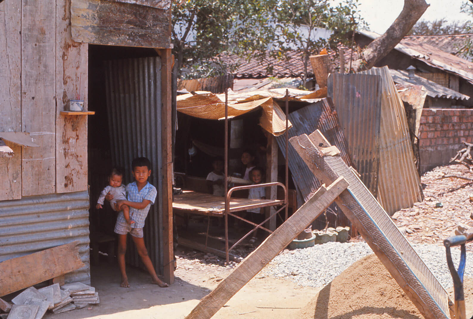 Young Vietnamese children standing in the doorways of temporary shelters constructed out of wood and corrugated steel.