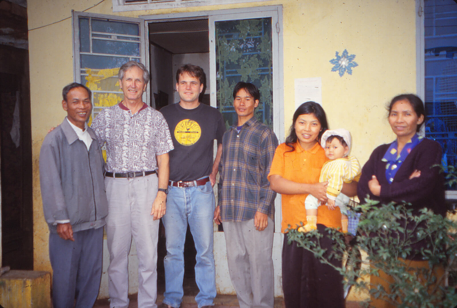 Two white men and an Asian family of 5 standing outside a yellow house.
