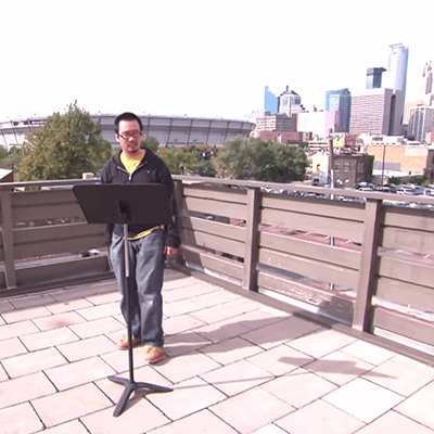 Contemporary image of a Vietnamese man standing on a rooftop in downtown Minneapolis, reading from a music stand.