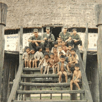 Soldiers sitting on steps up to a house with a group of Asian children.