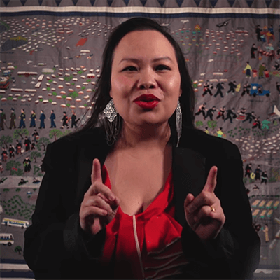 Contemporary image of a young Hmong woman gesturing with her two hands, standing in front of a Hmong story cloth.