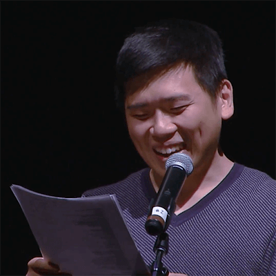 Contemporary image of a young Hmong man smiling at a microphone, reading from a sheet of paper.