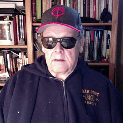 Contemporary image of an older gentleman in a "War Poet 69" zip up hoodie, a MN Twins ball cap, and sun glasses, sitting in front of shelves of books.