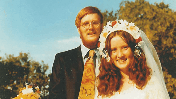 A young couple on their wedding day. The man wears a suit and glasses, the woman wears a floral crown.