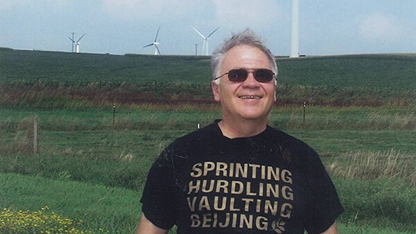 Contemporary photo of an older gentleman standing in a field of wind turbines, wearing sunglasses and a big smile.