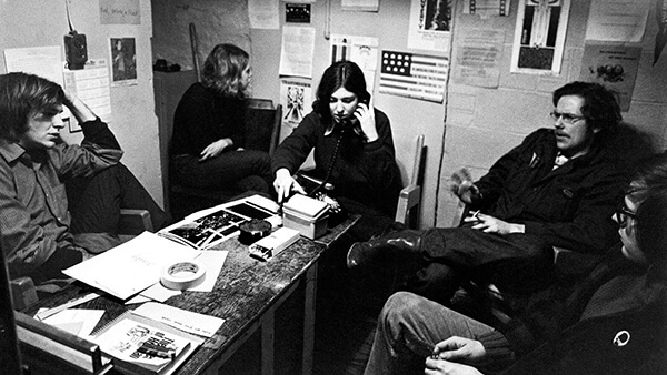 A woman at a desk, on the telephone, with three men seated around.