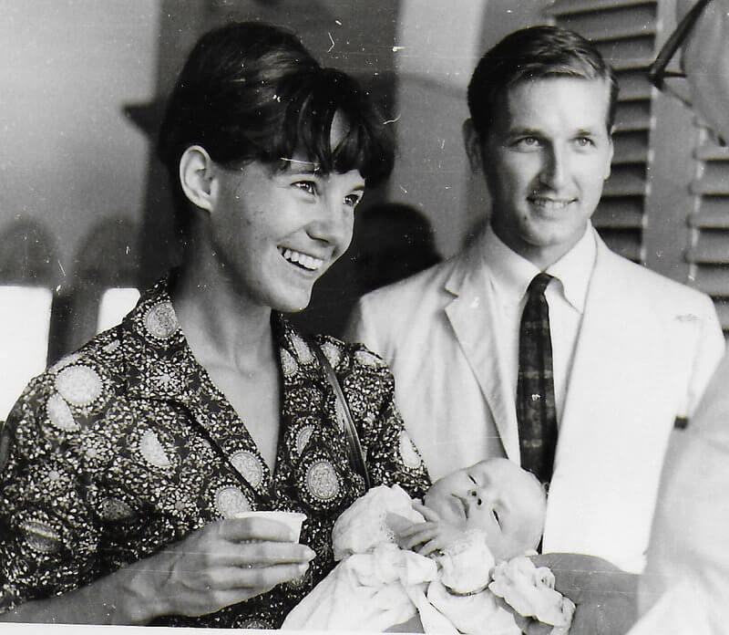 A black and white portrait of two beaming parents holding their infant and looking at a priest off camera.
