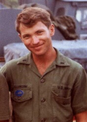A young man in green navy fatigues smiling toward the camera.