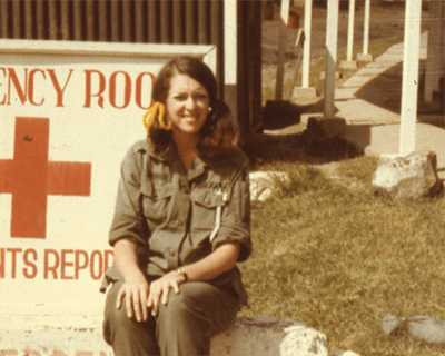 A young nurse in uniform and pigtails, sitting outside in front of an Emergency Room sign.