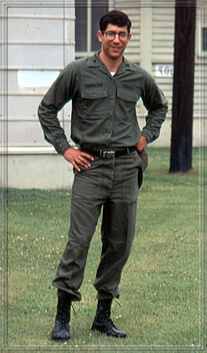 A bespectacled soldier standing outside a building with his hands on his hips.