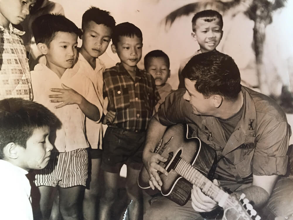 A soldier sitting with a guitar while Asian children gather around and look at him.