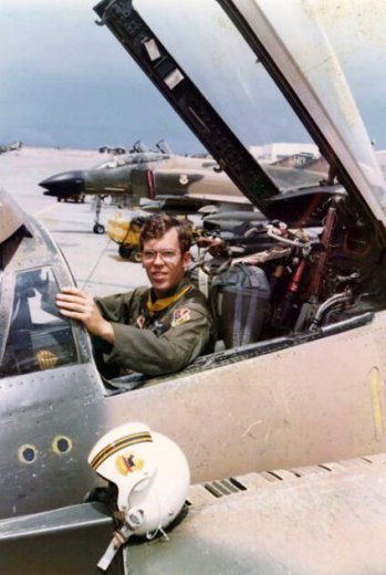 A young pilot posing in a cockpit of a fighter jet, helmet sitting nearby on the wing.