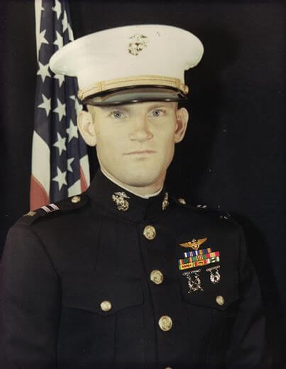 Official military portrait of a young Marine in dress blues, in front of an American flag.