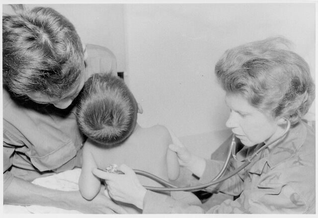 Two nurses checking on a small child. One holds him, and the other holds a stethoscope to his back.