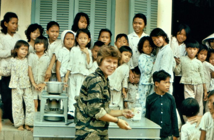 A young nurse in fatigues, sterilizing needles. A large group of Asian children is lined up in the background.