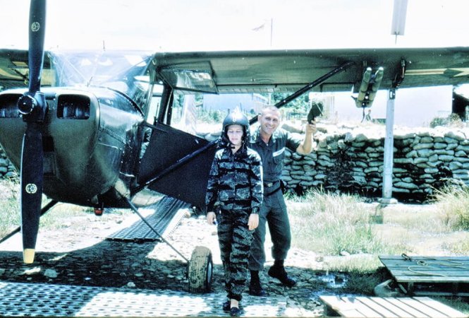 A young woman in fatigues and a pilots helmet, standing in front of a small plane while the pilot stands behind her and tips his hat at the camera.