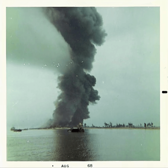 A large plume of dark smoke rising into the sky. Water and a small boat in the foreground; text on the photo margins reads "Aug 68".