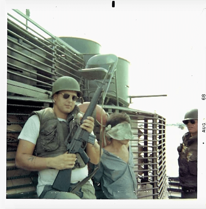 A young soldier wearing sunglasses and holding a rifle poses next to an Asian man who is blindfolded. Text on photo margins says "Aug 68".