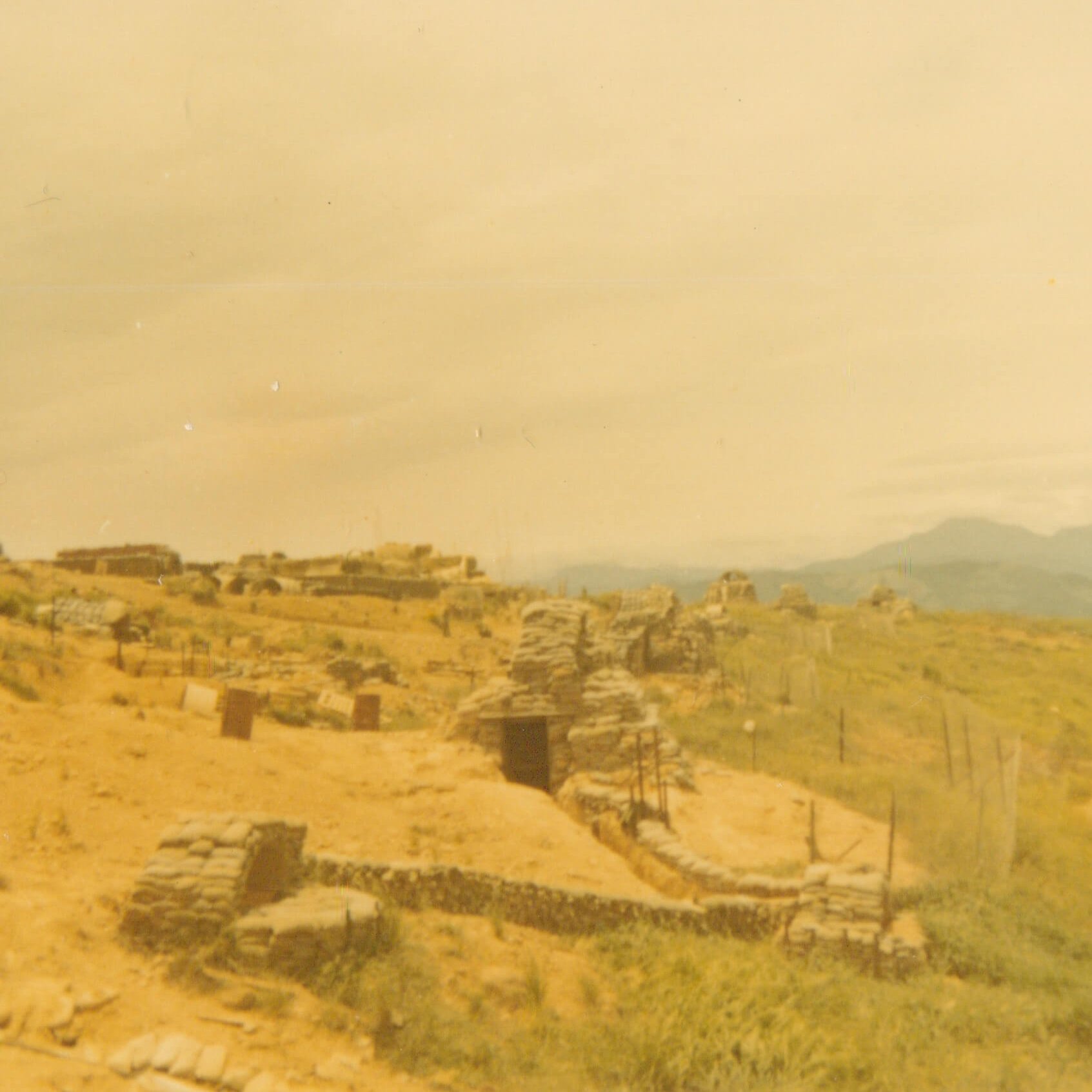 An encampment with sandbag structures and fencing; mountains are in the distance.
