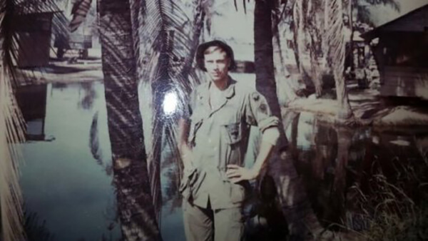 A young soldier with his hands on his hips, standing amid palm trees.