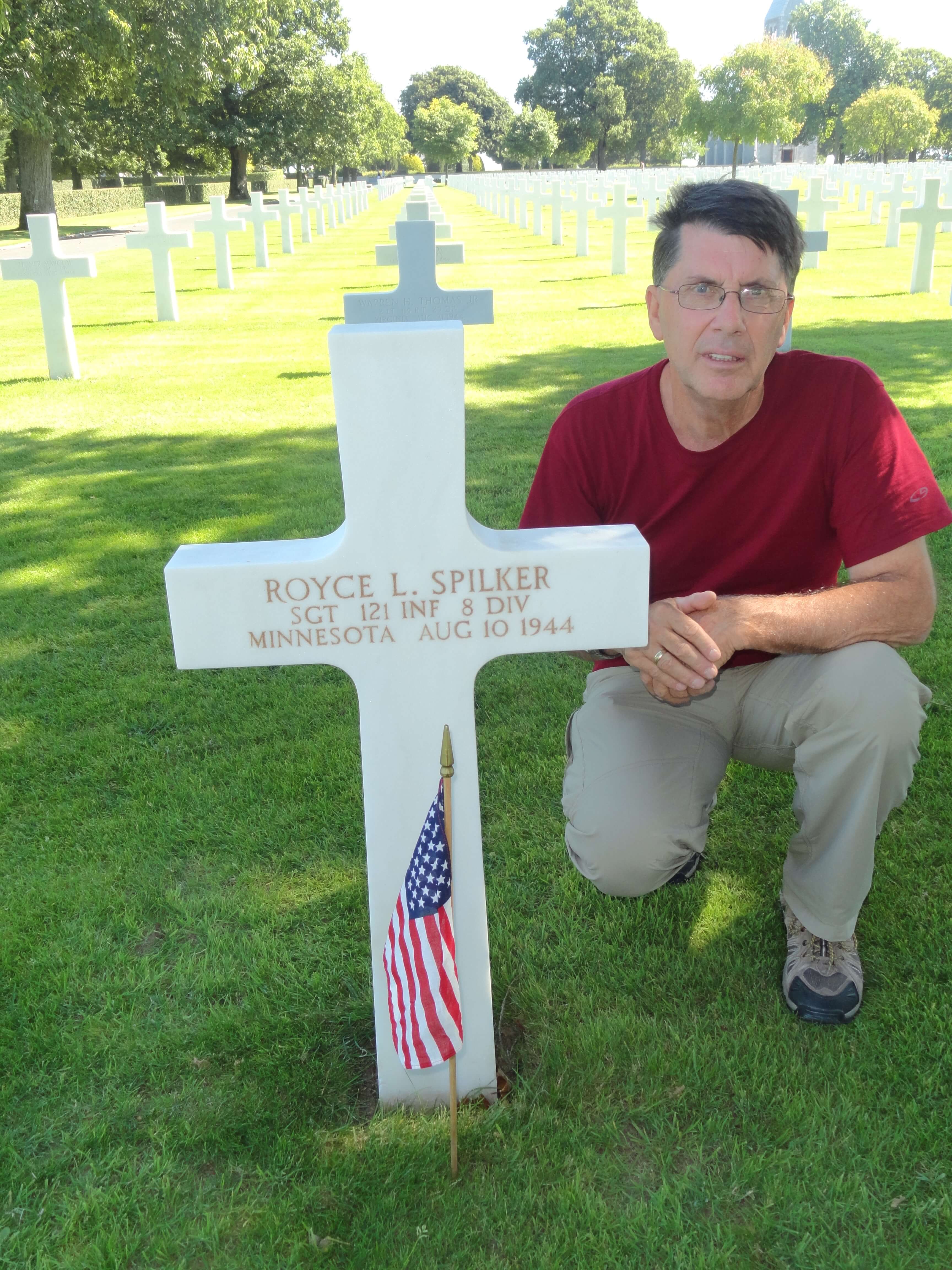 Contemporary photo of an older man squatting beside a cross grave marker for Royce L. Spilker.