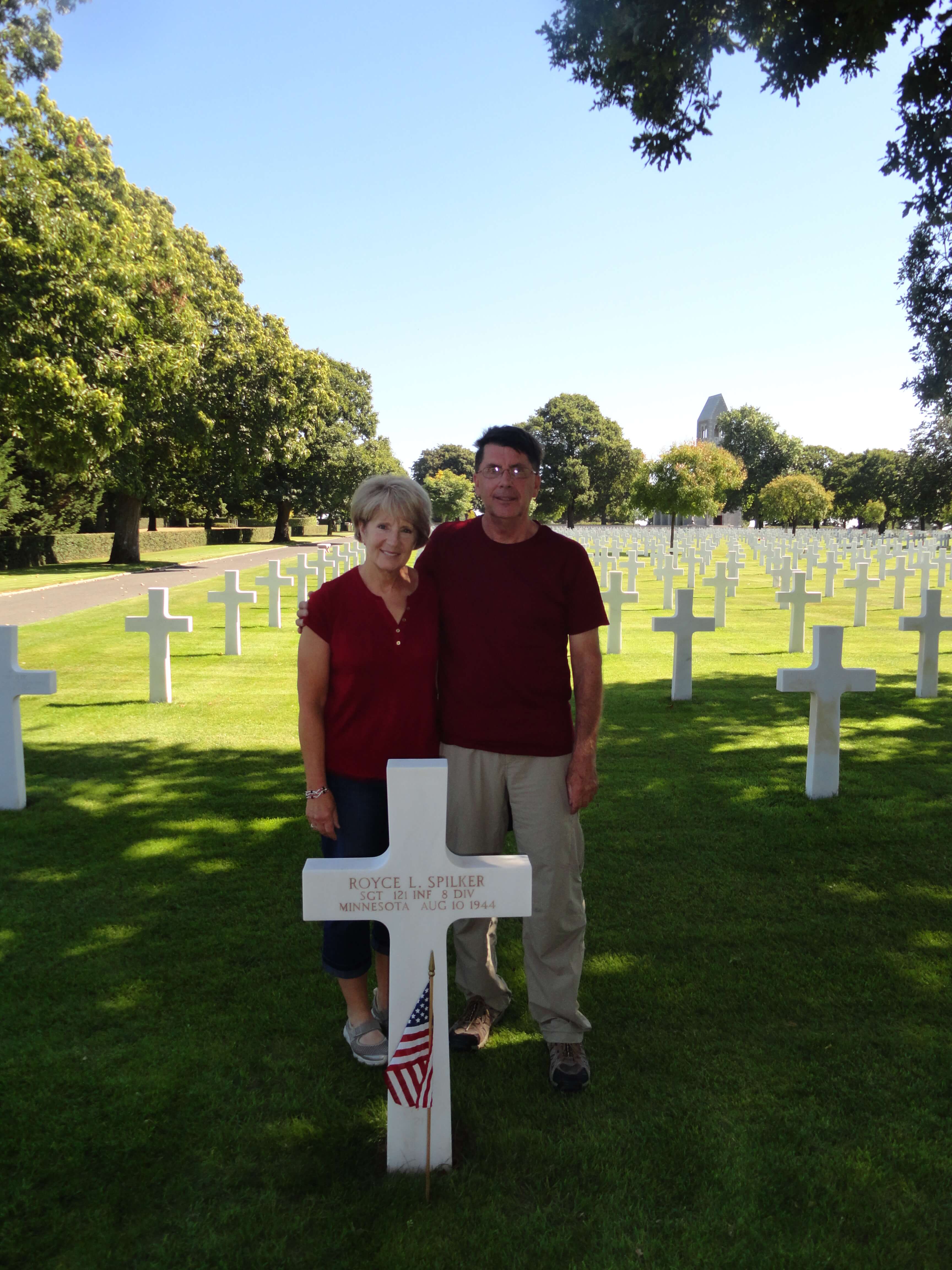 Contemporary photo of an older couple standing in a military cemetery behind a cross grave marker.